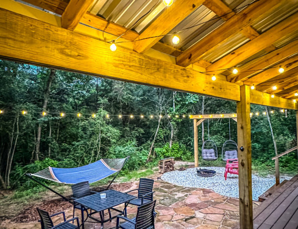 Patio of 1962 Airstream glamping Texas featuring a dining table with four chairs, a double hammock swing, and a fire pit surrounded by Adirondack chairs